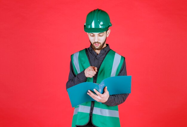 Engineer in green uniform and helmet holding a blue folder, reading and making corrections. 