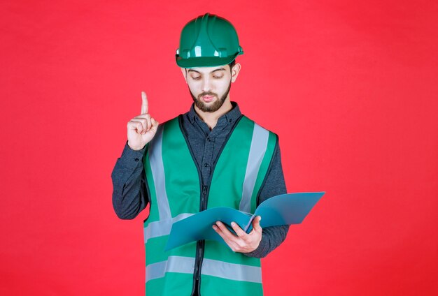 Engineer in green uniform and helmet holding a blue folder, reading and making corrections. 
