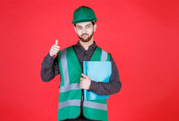 Engineer in green uniform and helmet holding a blue folder and showing positive hand sign. 