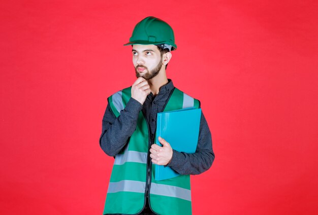 Engineer in green uniform and helmet holding a blue folder and thinking. 