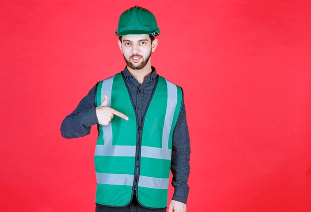 Engineer in green uniform and helmet pointing at herself. 