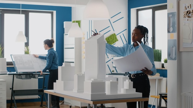 Engineer holding blueprints plan and analyzing building model for architecture work. Woman architect working with tools to design construction layout and structure for urban project.