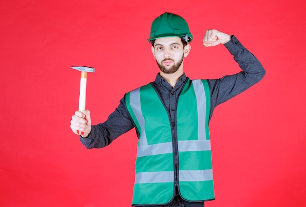 Бесплатное фото Инженер в зеленой форме и шлеме держит деревянный топор и показывает положительный знак рукой.
