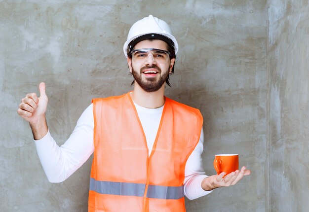 오렌지 머그잔을 들고 음료의 맛을 즐기는 흰색 헬멧에 엔지니어 남자.