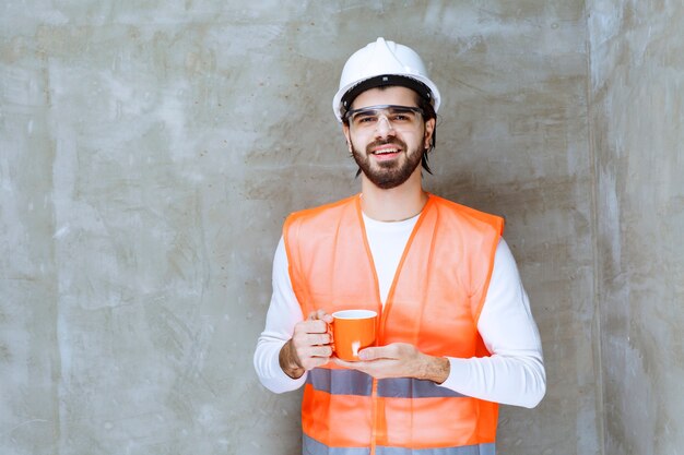 Инженер в белом шлеме держит оранжевую кружку.