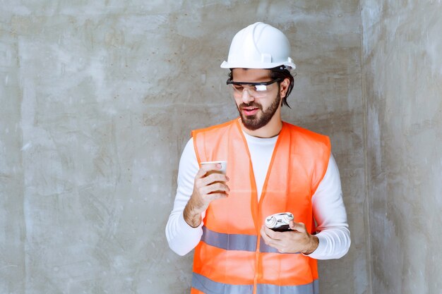 Engineer man in white helmet and protective eyeglasses holding an alarm clock and a cup of drink.