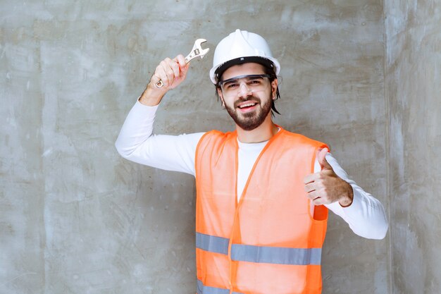 Инженер в белом шлеме и защитных очках держит металлические ключи и показывает знак удовольствия.