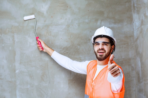 Free photo engineer man in white helmet and protective eyeglasses painting the wall with trim roller and showing thumb up.