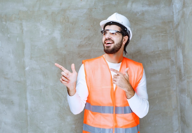 Engineer man in white helmet and protective eyeglasses pointing at his colleague or at something aside.