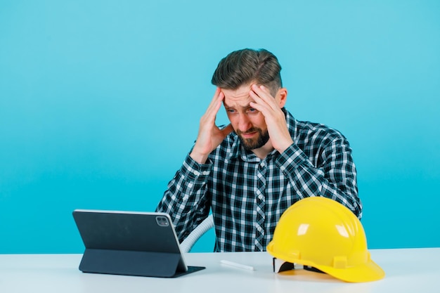 Engineer man with headache is holding hands on forehead on blue background