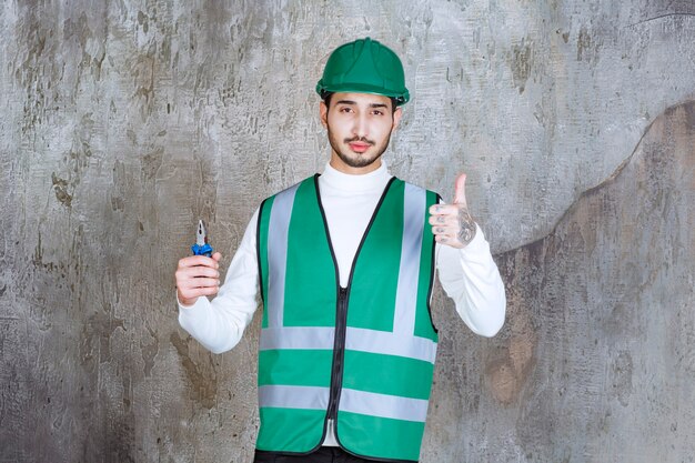 Engineer man in yellow uniform and helmet holding blue pliers for repair and showing positive hand sign.