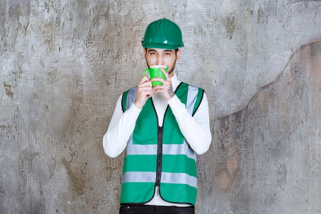 Free photo engineer man in yellow uniform and helmet holding a green coffee mug and smelling the product.