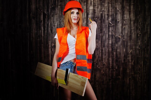 オレンジ色のエンジニアの女性は、ボードと定規を保持している木製の背景からヘルメットと建物のジャケットを保護します
