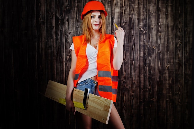 주황색의 엔지니어 여성은 보드와 통치자를 들고 나무 배경에 대해 헬멧과 건물 재킷을 보호합니다
