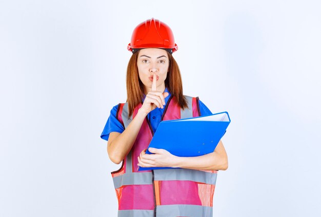 Engineer woman in red helmet holding a blue project folder and asking for silence. 