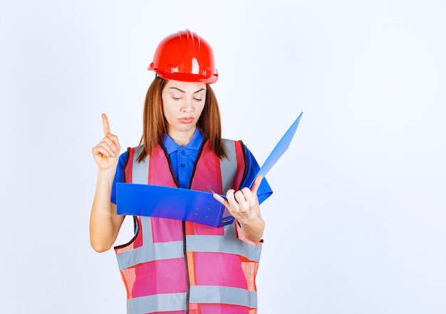 Free photo engineer woman in red helmet holding a blue project folder and checking the reports, raising finger to make corrections.