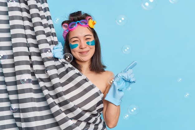 Free photo enjoy simple things. delighted brunette asian woman applies collagen pads under eyes hides naked body behind shower curtain indicates aside on blue background with bubbles has healthy soft skin