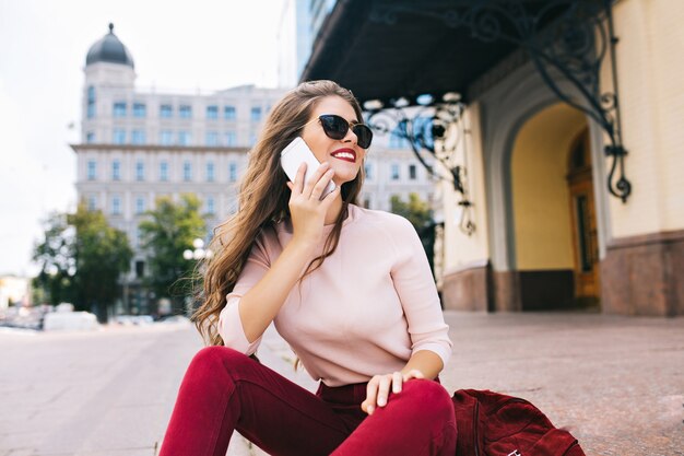 Понравившаяся девушка с длинной прической отдыхает на лестнице в городе. Она носит бордовые штаны, разговаривает по телефону и улыбается в сторону.