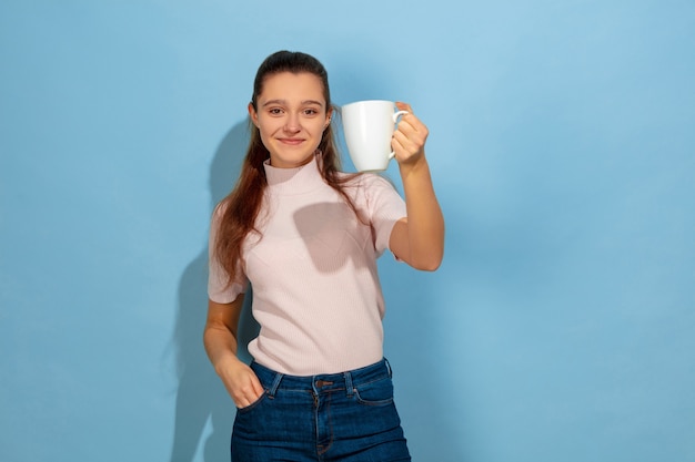 Пьет кофе, чай, выглядит спокойно. Портрет кавказской девочки-подростка на синем фоне. Красивая модель в повседневной одежде. Понятие человеческих эмоций, выражения лица, продаж, рекламы. Copyspace. Выглядит счастливым.