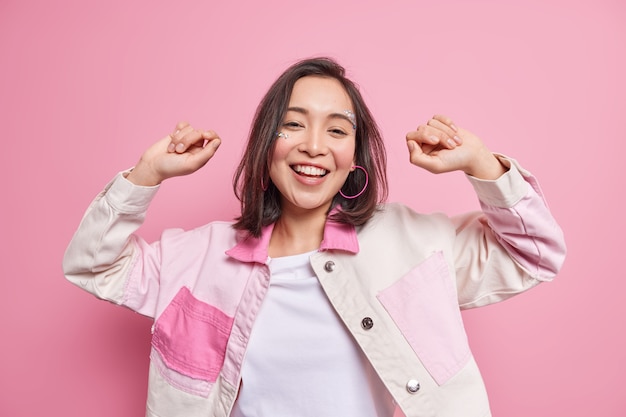 Enjoying life. Positive beautiful Asian millennial girl has fun and dances carefree smiles broadly dressed in stylish jacket isolated over pink wall moves to favorite song keeps hands up