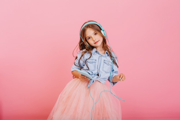 Free photo enjoying lovely music through blue head[hones of cute little girl with long brunette hair isolated on pink background. fashionable child in tulle skirt expressing true positive emotions to camera