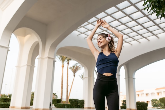 Enjoying lovely music through headphones of positive joyful sportswoman stretching hands above on east architecture. Fashionable model, having fun..