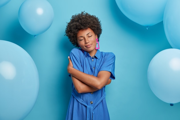 Enjoying pleasant moment. Charming curly haired woman embraces herself and stands with closed eyes, wears fashionable dress and pink earrings, remembers something positive, ready for celebration