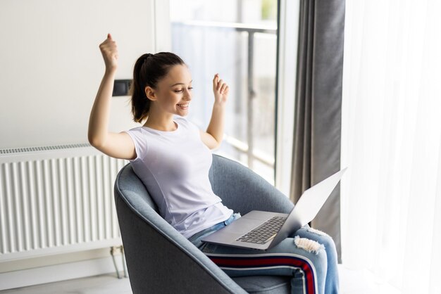 Enjoying relaxing time after work at home. Beautiful young smiling woman working on laptop and drinking coffee while sitting in a big comfortable chair at home.