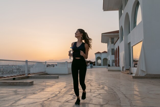 水のボトルを実行している美しい陽気な若い女性の海岸で夏の晴れた朝を楽しんでいます。目を閉じて側に笑顔、東熱帯の国、トレーニング、外でのトレーニング