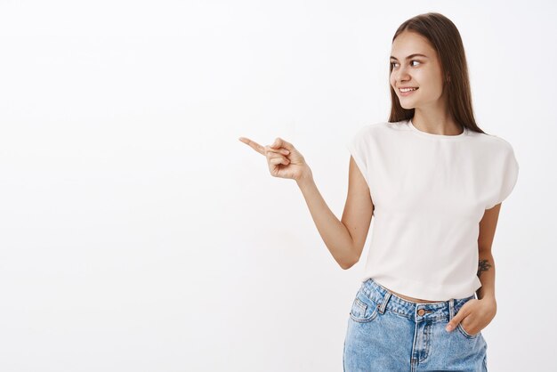 Entertained curious attractive and stylish caucasian woman in white blouse holding hand in jeans pocket turning and pointing left with interest smiling happily while standing over gray wall