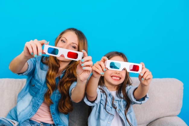 Entertainment, family time together of amazing mother with her young daughter on couch isolated on blue background. Showing 3D glasses to camera, smiling, expressing true happy emotions