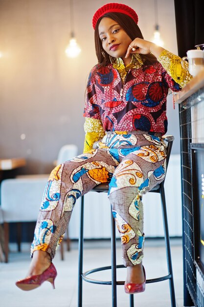 Восторженная африканская американка в модном цветном наряде с красным беретом отдыхает в уютном кафе, сидя на барной стойке