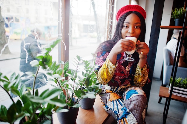 Восторженная африканская американка в модном цветном наряде с красным беретом отдыхает в уютном кафе с чашкой капучино в руках