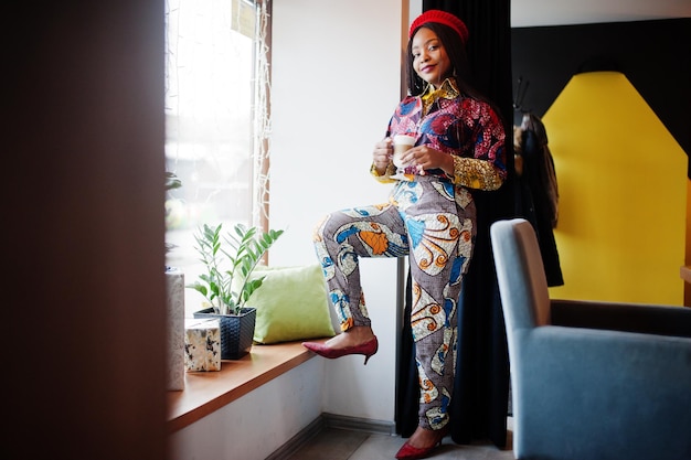 Enthusiastic african american woman in trendy coloured outfit with red beret chilling in cozy cafe with cup of hot latte in hands