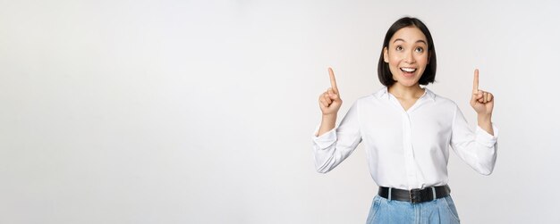 Enthusiastic asian business woman pointing looking up with happy smiling face showing company logo or banner standing over white background