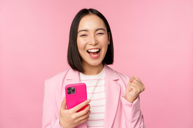 Enthusiastic asian businesswoman saying yes winning on mobile phone using smartphone and triumphing celebrating success standing over pink background