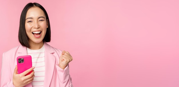 Enthusiastic asian businesswoman saying yes winning on mobile phone using smartphone and triumphing celebrating success standing over pink background
