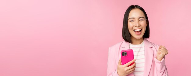 Enthusiastic asian businesswoman saying yes winning on mobile phone using smartphone and triumphing celebrating success standing over pink background