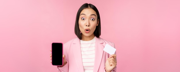 Enthusiastic asian businesswoman showing mobile phone screen and credit card looking amazed at camera standing over pink background