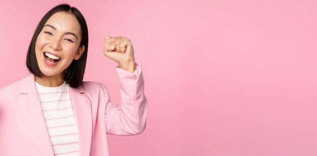 Enthusiastic asian corporate woman businesswoman raising hand up and cheering triumphing winning and celebrating standing over pink background