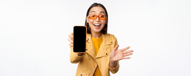 흰색 배경 위에 서 있는 휴대폰 화면에 스마트폰 앱 인터페이스 온라인 스토어 또는 웹사이트를 보여주는 열성적인 아시아 여성 모델