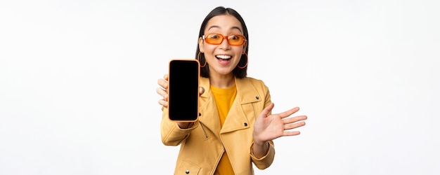 Enthusiastic asian female model showing smartphone app interface online store or website on mobile phone screen standing over white background