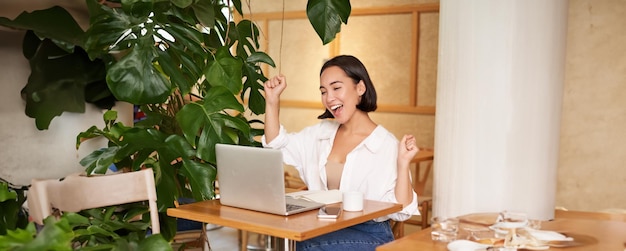 Free photo enthusiastic asian girl celebrating looking at laptop and triumphing makes fist pumps with excited