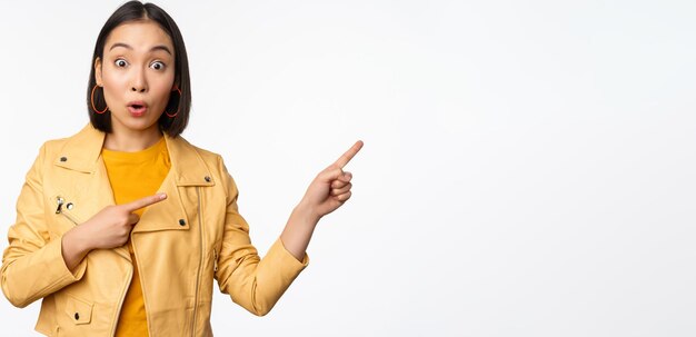 Enthusiastic asian girl pointing fingers right showing advertisement and smiling inviting to the store standing over white background