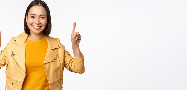 Enthusiastic asian girl pointing fingers up showing advertisement on top smiling happy demonstrating promo offer or banner standing over white background