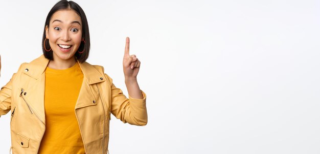 Восторженная азиатская девушка, указывающая пальцем вверх, показывает рекламу сверху, улыбаясь, счастливо демонстрируя промо-предложение или баннер, стоящий на белом фоне