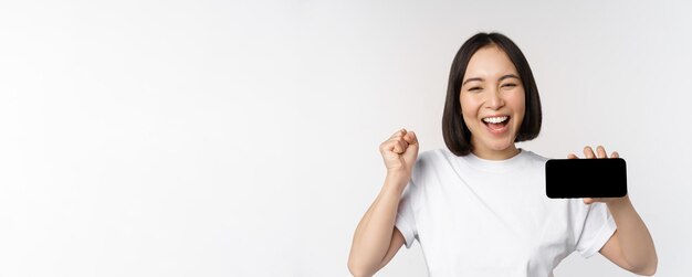 Enthusiastic asian girl scream in joy showing horizontal smartphone screen mobile phone display stan