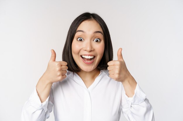 Enthusiastic asian girl showing thumbs up and smiling pleased by something recommending smth good standing over white background