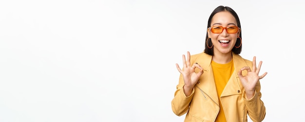 Enthusiastic asian girl in sunglasses shows okay ok sign in approval smiling and laughing approve recommend smth praise and compliment standing over white background