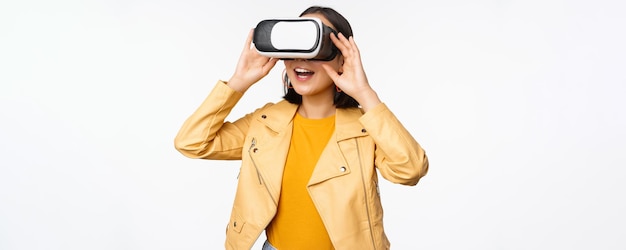 Enthusiastic asian girl in VR glasses looking with virtual reality headset at smth amazing standing over white background